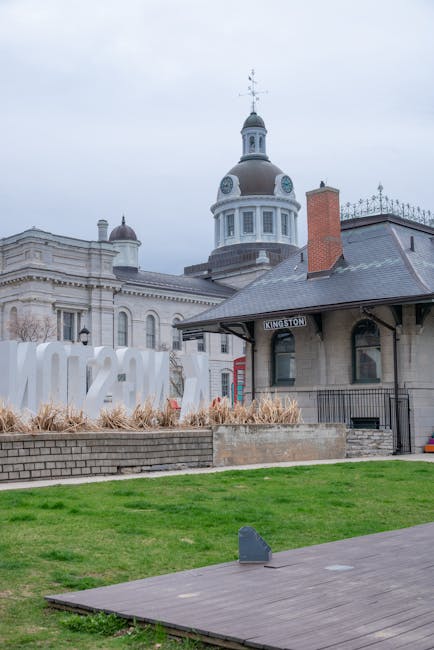 The image depicts a historic railway station building with a prominent dome and clock tower situated in an urban setting. The station's architecture features a light-colored façade with classical des