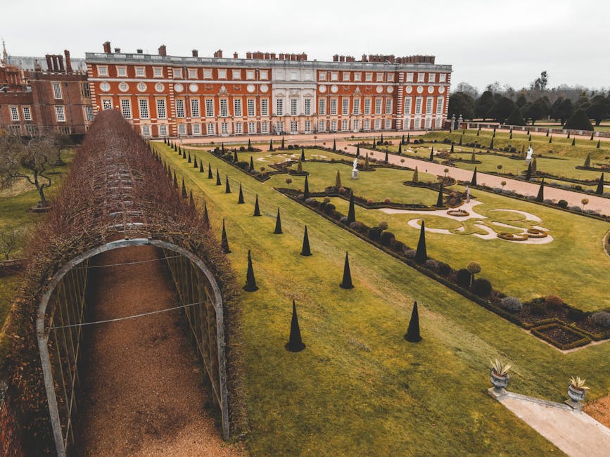 Aerial view of a large historic castle surrounded by extensive formal gardens and lush green lawns, with pathways and trimmed hedges arranged in symmetrical patterns. The castle features numerous bric